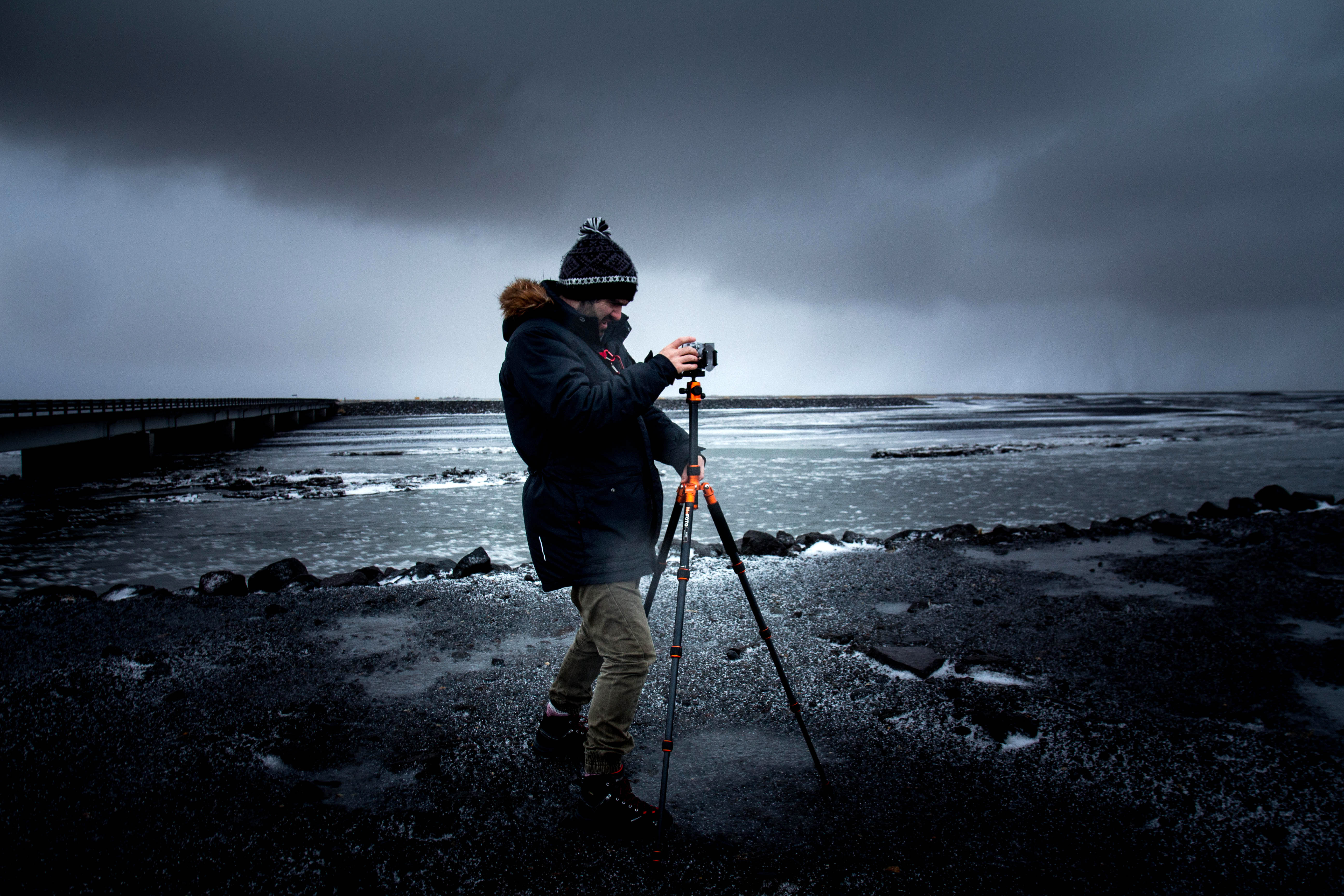 manincoatandhatsettinguptripodonbeachunderclouds image 