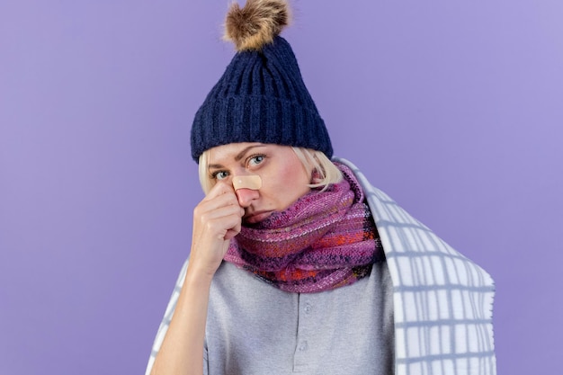 Sad young blonde ill slavic woman with medical plaster on nose wearing winter hat and scarf