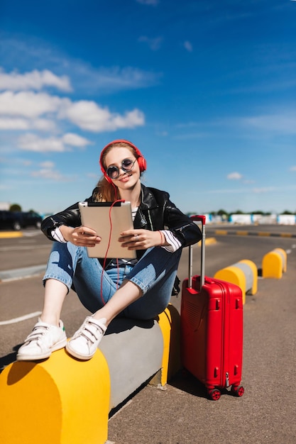 Smiling girl in sunglasses and headphones listening to music on tablet with red suitcase near airport road