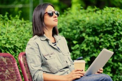 Young Attractive Brunette Female in Sunglasses Holding Tablet PC and Drinking Coffee in Green Summer Park