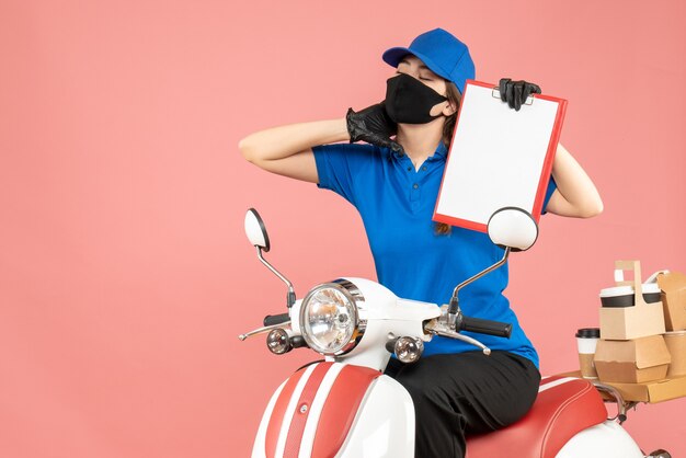 Front view of overtired courier woman wearing medical mask and gloves sitting on scooter holding empty paper sheets delivering orders on pastel peach background