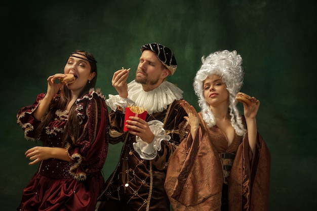 Delicious fastfood. Portrait of medieval young people in vintage clothing on dark background