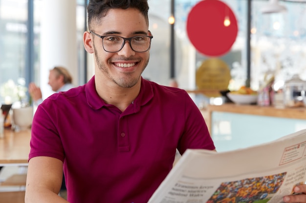 Glad hipster guy with glasses reading good news in newspaper in cafeteria