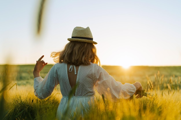 Young beautiful woman with blond long hair in a white dress in a straw hat collects flowers on a wheat field. Flying hair in the sun, summer. Time for dreamers, golden sunset.