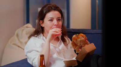 Business woman watching TV eating burger in living room after work. Young adult enjoying fast food delivery meal for dinner while sitting on sofa and laughing at television screen