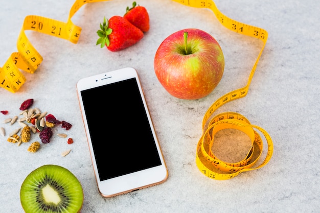 Dried fruits; apple; halved kiwi; strawberry; measuring tape and smartphone on gray textured background