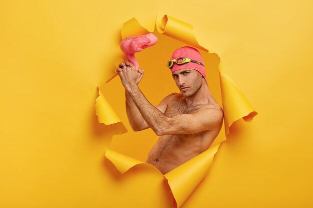 Self confident serious guy with bristle holds pink inflated flamingo, wears swimcap with goggles on forehead