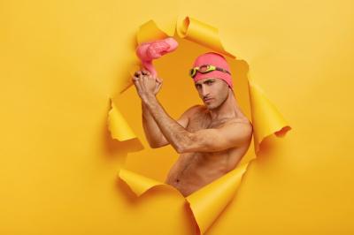 Self confident serious guy with bristle holds pink inflated flamingo, wears swimcap with goggles on forehead