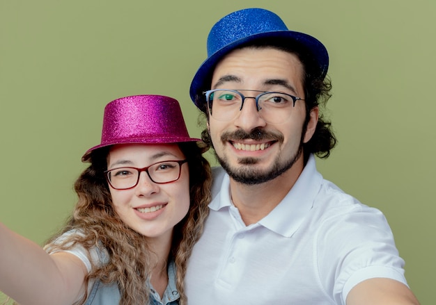 Smiling young couple wearing pink and blue hat holding camera isolated on olive green