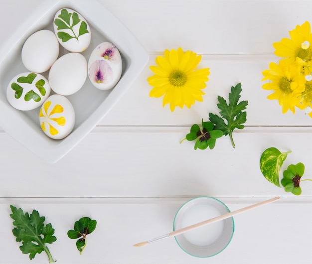 Easter Eggs in Container Surrounded by Flowers, Leaves, and Cup with Dye Liquid