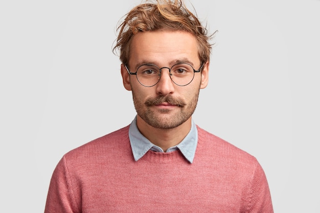 Serious man teacher with confident clever look, has beard and mustache, listens pupil`s answer, wears pink sweater, round glasses