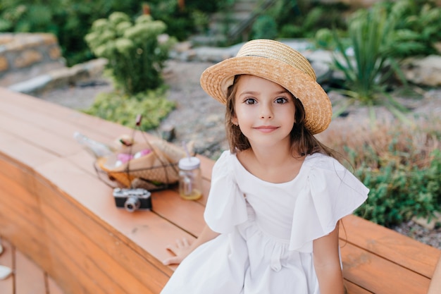 Outdoor Portrait of Dark-Eyed Girl in Vintage Straw Hat on Wooden Bench with Picnic Basket and Camera