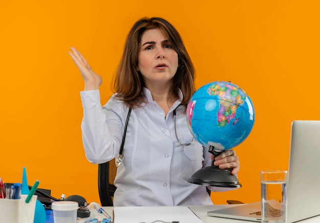 Middle-Aged Female Doctor Working at Desk with Medical Tools and Globe on Orange Wall