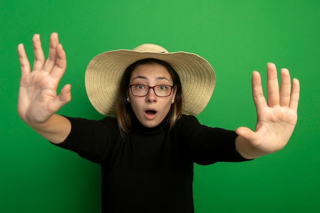 Young beautiful woman in summer hat and glasses making stop gesture over green wall