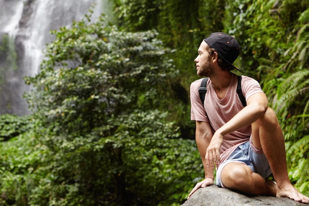 Travel and Adventure: Handsome Young Barefooted Male Hiker with Backpack Relaxing Alone on Big Stone and Looking Back