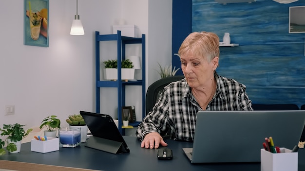 Mature Woman Using Tablet and Laptop to Analyze Financial Graphs While Working from Home