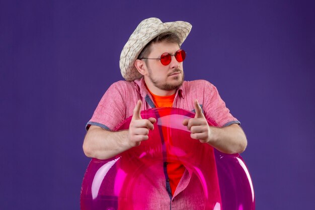 Young handsome traveler man in summer hat with inflatable ring pointing with fingers