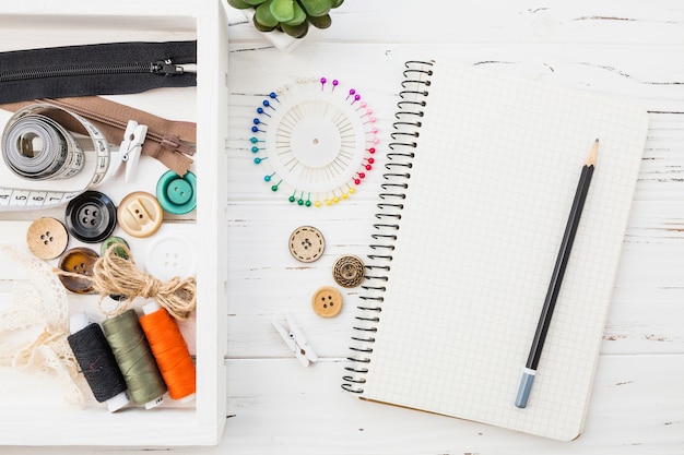 Various Sewing Accessories with Notepad and Pencil on Wooden Background