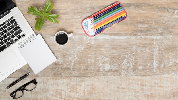 Wooden Desk Setup with Opened Laptop, Tea, Eye Glasses, Pen, Plant, and Colorful Pencils