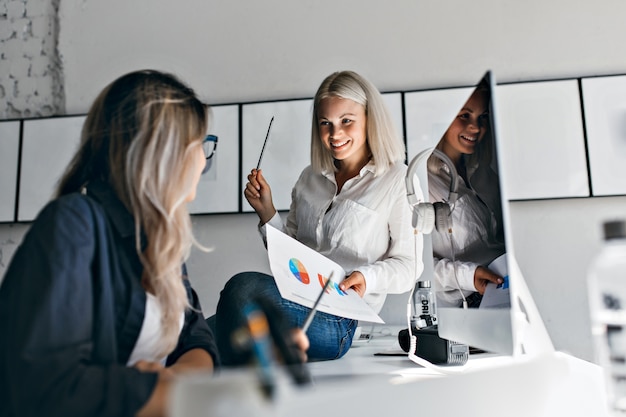 Smiling blonde female manager holding infographic and pencil, while sitting on table. Indoor portrait of two women working with computer in office