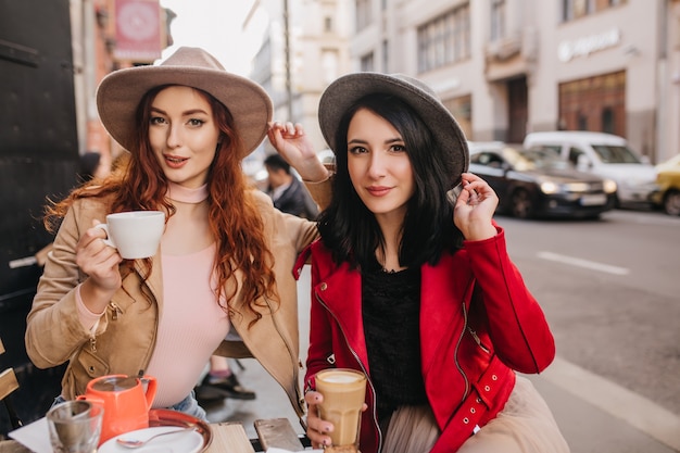 Brunette Woman in Gray Fedora Enjoying Time with Ginger Friend in CafÃ© – Free Download