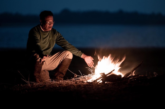 Senior Man Near Lake Warming Hands at Bonfire in Cold Morning – Free Download