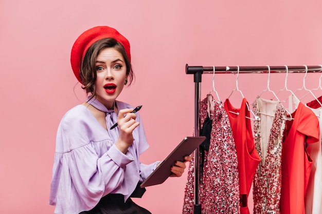 Lady with Green Eyes and Red Hat Holding Tablet Among Clothes Hangers – Free Stock Photo, Download for Free