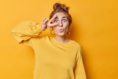 Teenage Girl Making Peace Gesture Over Eye with Silver Earrings on Yellow Background – Free Stock Photo