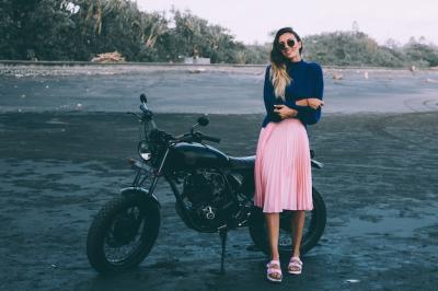 Stylish Woman in Sunglasses, Blue Sweater, and Pink Skirt by Black Bike on Black Sand Beach – Download Free Stock Photo