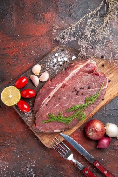 Overhead View of Red Meat with Garlic, Green Lemons, Onions, Fork, and Knife on a Dark Background – Free Download