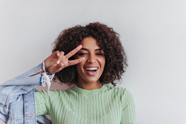 Brunette Girl Winking with Peace Sign – Happy Woman in Green Tee and Denim Outfit on White Backdrop | Free Download