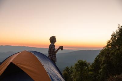 Happy Man Holding Coffee Cup Near Tent Amidst Mountains at Sunset – Free Download