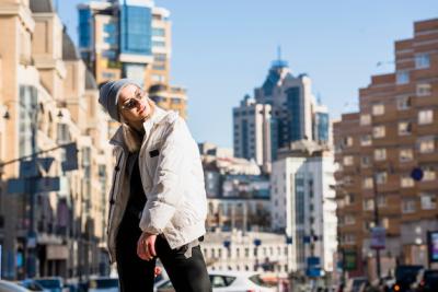 Attractive Smiling Young Woman in Glasses and Jacket Posing in Front of Buildings – Free Download