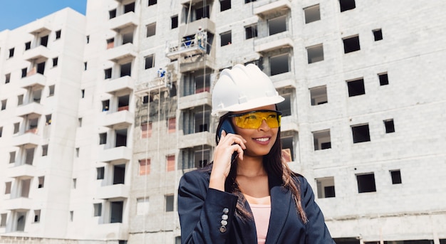 African American Lady in Safety Helmet Talking on Smartphone Near Construction Site – Free Download