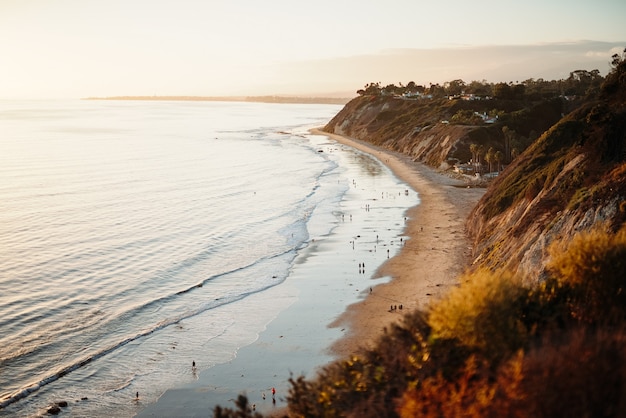 People Walking on a Scenic Seashore Next to Low Hills – Free Download