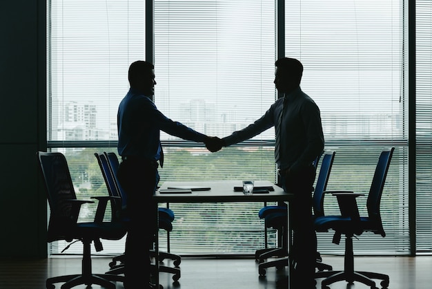 Silhouettes of Two Unrecognizable Men Shaking Hands in the Office – Free Download