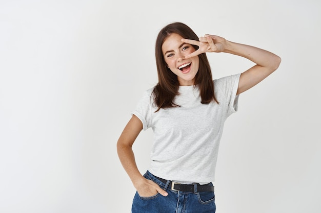 Brunette Woman Making Peace Sign and Smiling Against White Wall – Free Stock Photo for Download