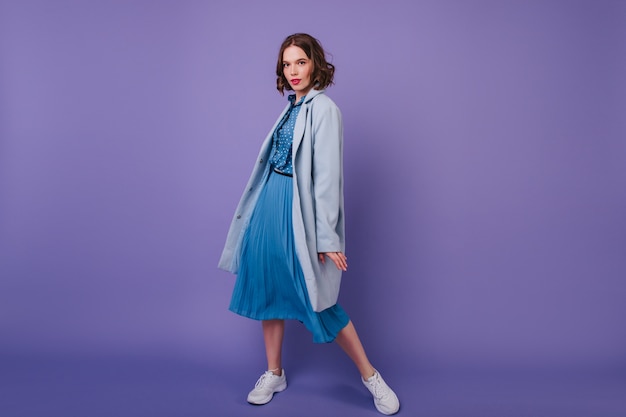 Refined Girl in Sneakers and Coat: Ecstatic Young Woman Posing in Blue Outfit against Purple Wall – Free Download