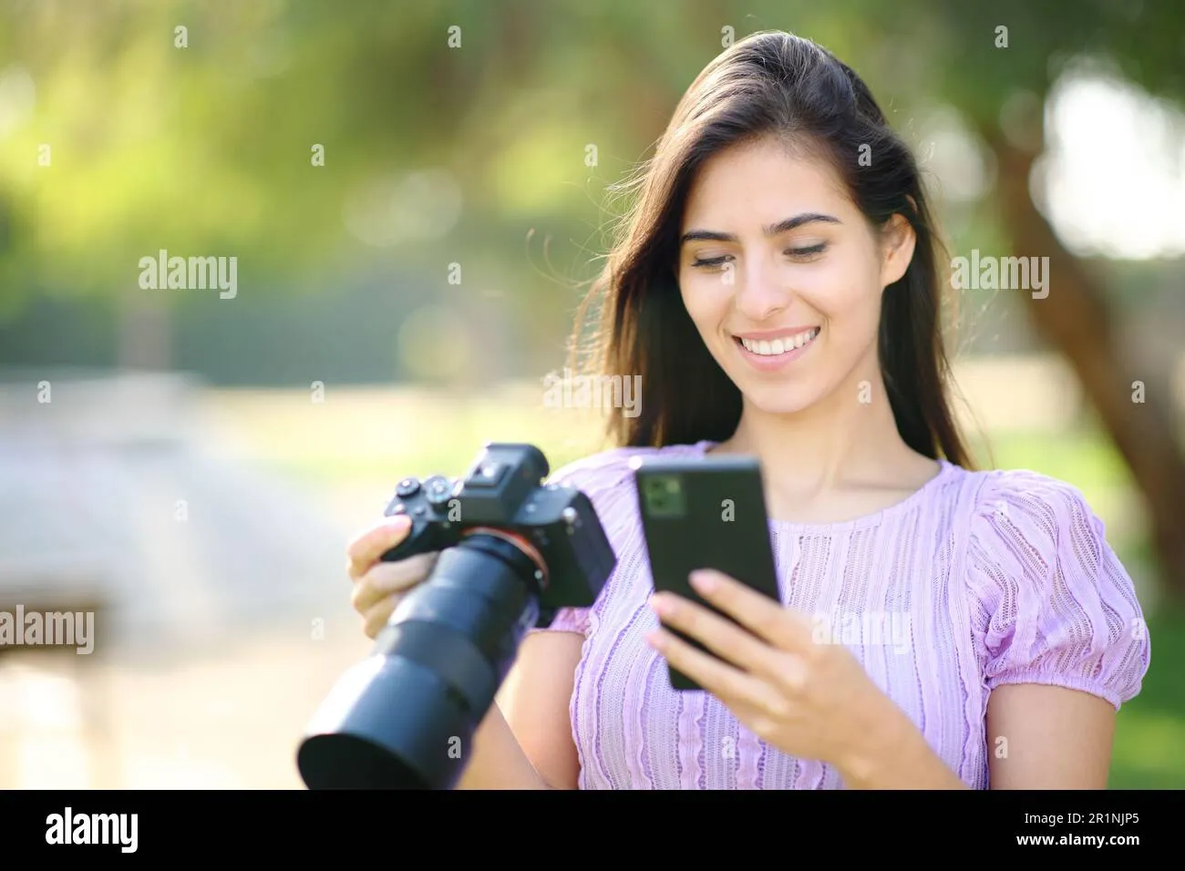 Happy photographer using phone and mirrorless camera in a park Stock 